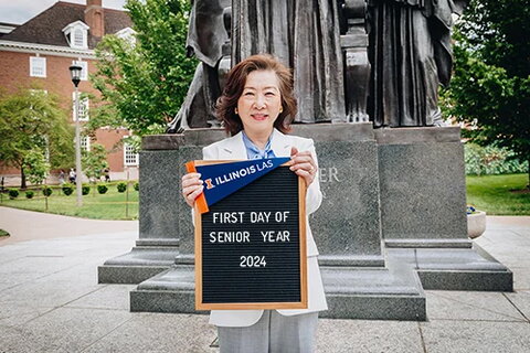 Bonnie Kim poses in front of Alma Mater with a sign saying "first day of senior year."